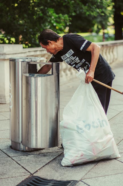 A man with dark hair, wearing a black T-shirt with white text, is seen disposing of waste into a metal public disposal bin with multiple separate openings, located on a paved walkway. He holds a large white plastic bag filled with rubbish in his left hand, while his right hand is positioned near the bin’s opening as he leans forward to deposit waste. The background features lush green trees and a stone balustrade, indicating an outdoor setting in a park or urban area. The scene appears to be during daylight with natural lighting and shadows cast on the pavement. This image depicts a private or independent waste disposal activity, which complements rubbish removal services by illustrating personal efforts to maintain cleanliness outside residential or public spaces. The polished metal surface of the bin reflects some of the surrounding greenery and daylight, highlighting the modern design typically seen in urban rubbish collection points, supporting the context of rubbish management and clearance.