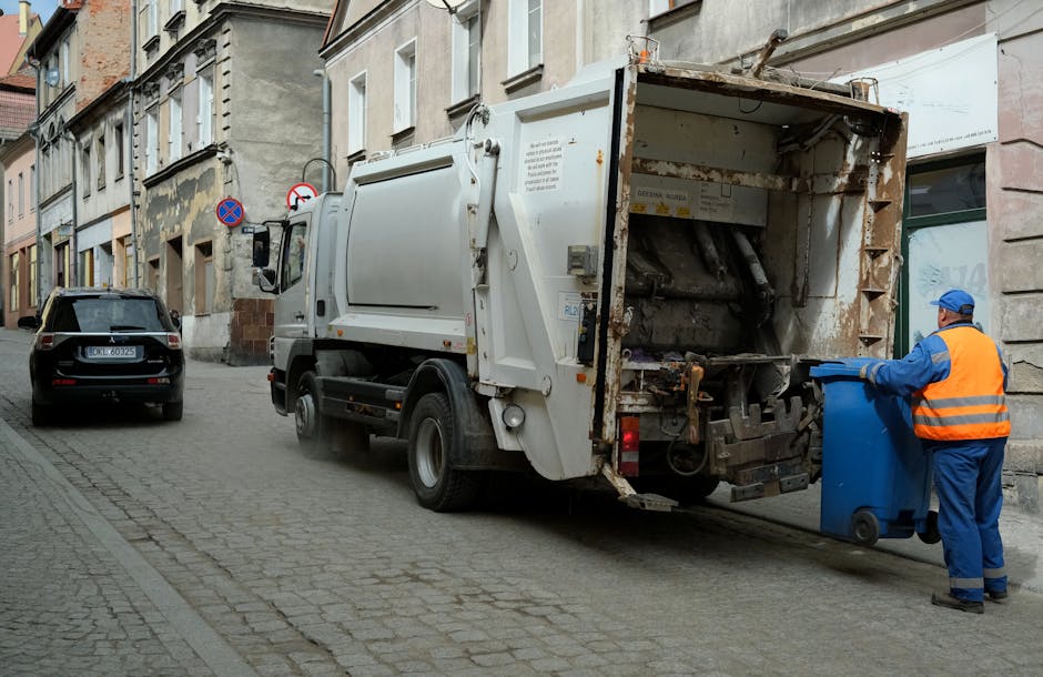 A street scene showing a large white rubbish collection truck parked alongside a row of old, multi-storey buildings with weathered facades. The truck's rear section is open, revealing an empty, rusted metal compactor designed for waste collection. On the right side of the image, a sanitation worker wearing a blue uniform, orange high-visibility vest, and matching cap is emptying a small blue wheeled bin into the truck's hopper. The street surface is paved with cobblestones, and a black SUV is parked further down the narrow lane. Visible street signs include a no-parking sign and a no-stopping sign mounted on a wall. The scene is lit by natural daylight, indicating daytime, with a generally neutral and professional tone reflective of waste collection operations, as carried out by services like Rubbish Clearance Hounslow that provide independent rubbish removal and disposal solutions in urban areas.