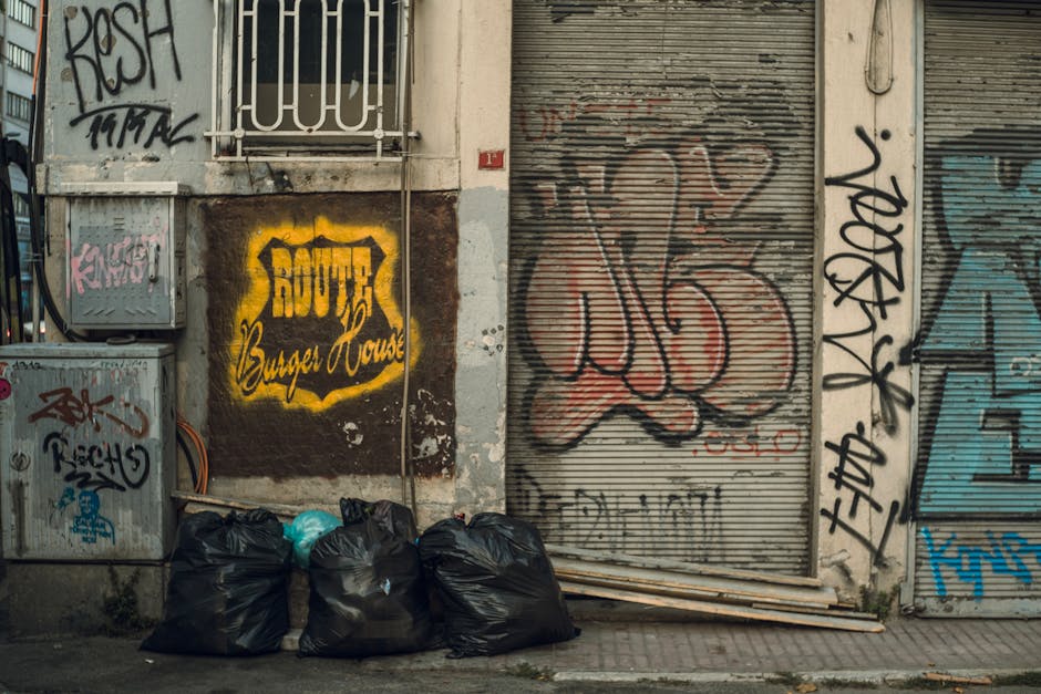 A man with dark hair, wearing a black T-shirt with white text, is seen disposing of waste into a metal public disposal bin with multiple separate openings, located on a paved walkway. He holds a large white plastic bag filled with rubbish in his left hand, while his right hand is positioned near the bin’s opening as he leans forward to deposit waste. The background features lush green trees and a stone balustrade, indicating an outdoor setting in a park or urban area. The scene appears to be during daylight with natural lighting and shadows cast on the pavement. This image depicts a private or independent waste disposal activity, which complements rubbish removal services by illustrating personal efforts to maintain cleanliness outside residential or public spaces. The polished metal surface of the bin reflects some of the surrounding greenery and daylight, highlighting the modern design typically seen in urban rubbish collection points, supporting the context of rubbish management and clearance.