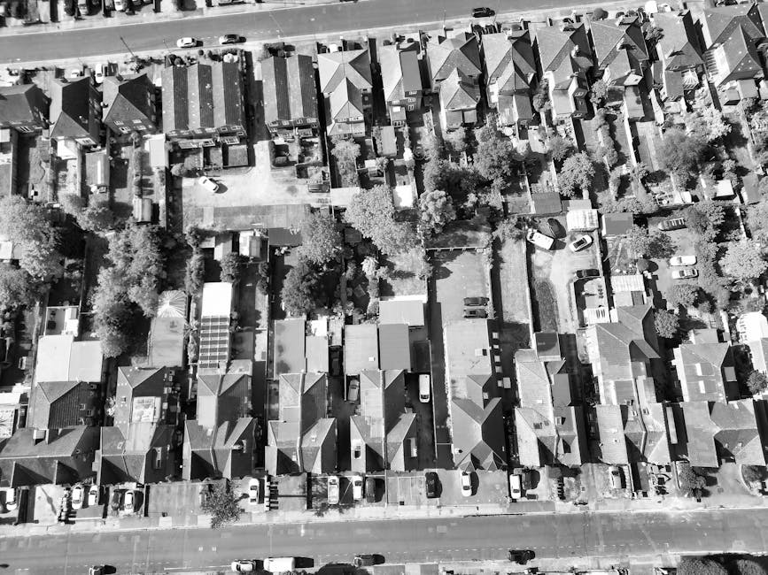 An aerial black-and-white photograph of a residential suburban area showing a row of terraced houses with pitched roofs, arranged in a uniform pattern along a straight street. Behind the row of houses, there are several backyards and small gardens separated by fences, with some yards containing trees, bushes, and outbuildings. Vehicles, mostly compact cars, are parked along the street and within the driveways of individual properties. The scene captures the density of the housing layout and the organized arrangement of vehicles typical of an urban neighbourhood. The overall environment appears quiet, with natural light illuminating the rooftops and street, emphasizing the textures of tile and slate roofs, as well as the paved surfaces. This image relates to private household waste management, illustrating the typical setting where rubbish removal services, such as those provided by Rubbish Clearance Hounslow, might operate for home clearance or on-site rubbish collection within local residential areas.