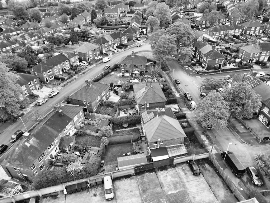 An aerial black-and-white photograph of a residential suburban area showing a row of terraced houses with pitched roofs, arranged in a uniform pattern along a straight street. Behind the row of houses, there are several backyards and small gardens separated by fences, with some yards containing trees, bushes, and outbuildings. Vehicles, mostly compact cars, are parked along the street and within the driveways of individual properties. The scene captures the density of the housing layout and the organized arrangement of vehicles typical of an urban neighbourhood. The overall environment appears quiet, with natural light illuminating the rooftops and street, emphasizing the textures of tile and slate roofs, as well as the paved surfaces. This image relates to private household waste management, illustrating the typical setting where rubbish removal services, such as those provided by Rubbish Clearance Hounslow, might operate for home clearance or on-site rubbish collection within local residential areas.