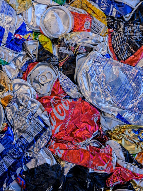 A close-up view of a pile of crushed and crumpled aluminum cans, primarily soda cans, with metallic surfaces showing various colors such as silver, red, blue, yellow, and black. The cans exhibit textures of smooth, glossy metal and ridges from tearing or crushing, with some featuring visible branding and logos, including a prominent red Coca-Cola can. The cans are stacked and jumbled together, with some partially overlapping and others leaning at different angles, creating a dense, chaotic arrangement. The background suggests an outdoor or industrial setting, as no specific environment is clearly visible. The scene illustrates typical waste materials collected for disposal or recycling, highlighting the materials handled by independent waste collection services like Rubbish Clearance Hounslow for rubbish removal or recycling. The lighting is even, emphasizing the metallic sheen and colorful labels on the cans, reflecting the importance of environmentally conscious disposal methods involving reuse and recycling.