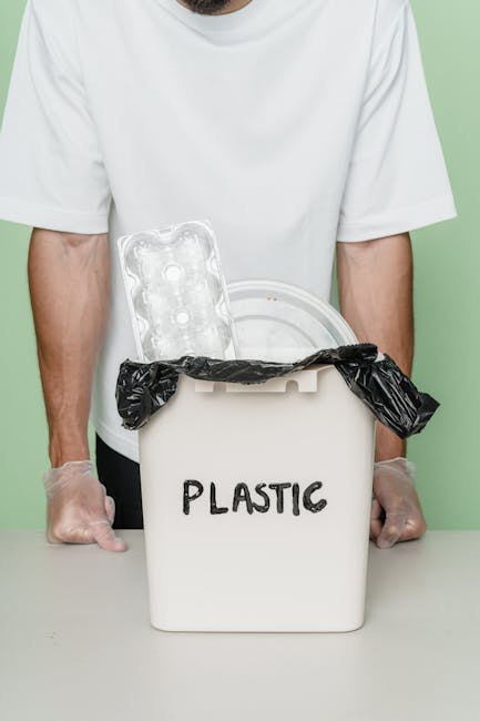 A person wearing a white t-shirt and transparent gloves is holding a white plastic bin labeled 'PLASTIC', which is partially lined with a black plastic bag. The open bin contains a clear plastic egg carton and a transparent plastic lid. The scene is set against a plain, light-colored background, possibly green or pastel, with the individual standing behind a flat surface. The image clearly depicts household plastic waste ready for collection or disposal, highlighting the importance of proper rubbish handling. The focus is on the process of sorting and preparing plastics for recycling or alternative waste handling, aligning with eco-conscious rubbish removal practices such as those offered by Rubbish Clearance Hounslow.