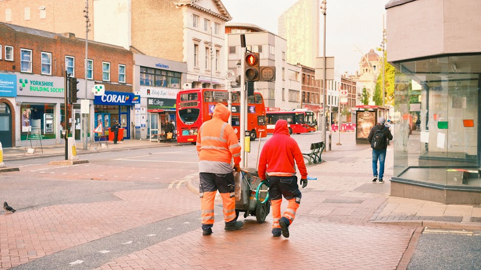 Two workers dressed in orange high-visibility uniforms are walking along a city street, pushing a trolley loaded with waste containers. They are positioned in the foreground, with their backs to the camera. One worker holds the handle of the trolley, while the other guides it. The street features a mix of commercial buildings, including a store with a green sign for the Yorkshire Building Society and another with a Coronation-branded advertising display. In the background, a red double-decker bus is stopped at a traffic light, which displays a red signal. Other pedestrians are walking along the pavement, and a bus stop shelter with a glass front is visible on the right side of the image. The environment is urban with a typical city centre atmosphere, under early morning or late afternoon lighting that provides a warm tone. This scene appears to be part of an independent waste collection or rubbish clearance operation, suitable for a professional rubbish removal service like Rubbish Clearance Hounslow, indicating efforts to maintain or clear the area of waste material.