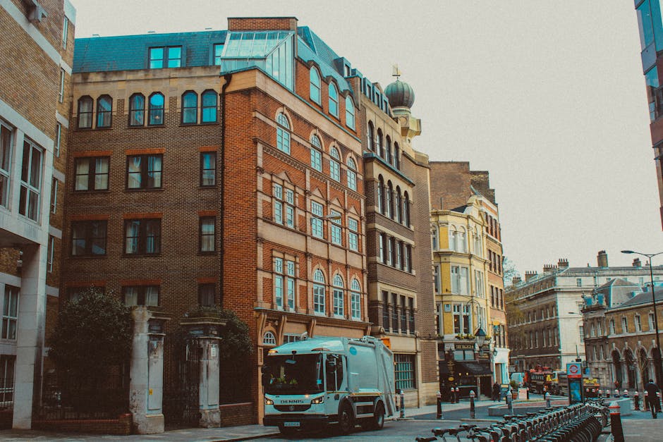 A narrow city street flanked by tall, multi-story buildings featuring a mix of brick and ornate architectural details, with large windows and decorative elements. In the foreground, a blue waste collection vehicle is parked on the paved road, positioned near the curb, with a small section of sidewalk visible behind it. To the right, several metal bike racks are aligned along the pavement. Further down the street, a few vehicles and buildings with classical facades can be seen, along with streetlights and a sign indicating a bus stop. The scene is illuminated by natural daylight, with an overcast sky contributing to diffuse, even lighting. This urban setting reflects a typical central London environment, where private waste collection services like Rubbish Clearance Hounslow might operate to manage rubbish removal in a busy commercial and residential area, highlighting the contrast between historic architecture and modern waste management solutions.