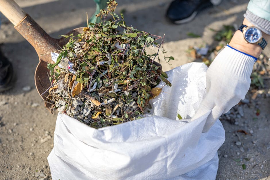 A person wearing a white glove is holding a white sack filled with garden waste, including a mix of small branches, leaves, and twigs, placed on a gravel surface. The person’s wrist features a silver watch, and a small metal shovel with a wooden handle is partially visible, resting against the sack and containing additional plant debris. The background shows a paved area with some blurred leaves and scattered litter, indicating an outdoor setting likely in a garden or yard. The scene reflects an on-site clearance activity where garden waste is being prepared for removal by a professional rubbish collection service, such as Rubbish Clearance Hounslow, supporting alternative, private waste handling options outside local authority collection.