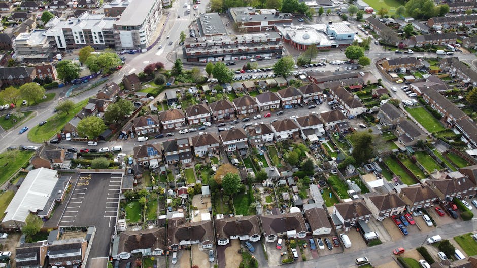 An aerial view of a residential suburban area showing multiple rows of terraced houses with small back gardens, many of which contain patio furniture, sheds, and garden ornaments. The houses are constructed with red brick and feature tiled roofs, with some properties having solar panels installed on the roofs. The back gardens are fenced and display a variety of greenery, including trees, bushes, and lawns. Alongside the houses, a narrow street runs through the area, lined with parked cars of different makes and colors. To the left of the image, there is a large, flat-roofed building with an adjacent parking lot filled with vehicles, possibly a community centre or commercial premises. In the background, a mixture of commercial buildings and larger residential complexes can be seen, with trees and greenery interspersed across the scene. The overall environment appears well-maintained, with natural daylight illuminating the scene, suggesting a clear and mild day. This setting illustrates typical on-site domestic waste management, with potential for private rubbish clearance services like those offered by Rubbish Clearance Hounslow, as part of alternative waste handling methods for household rubbish.