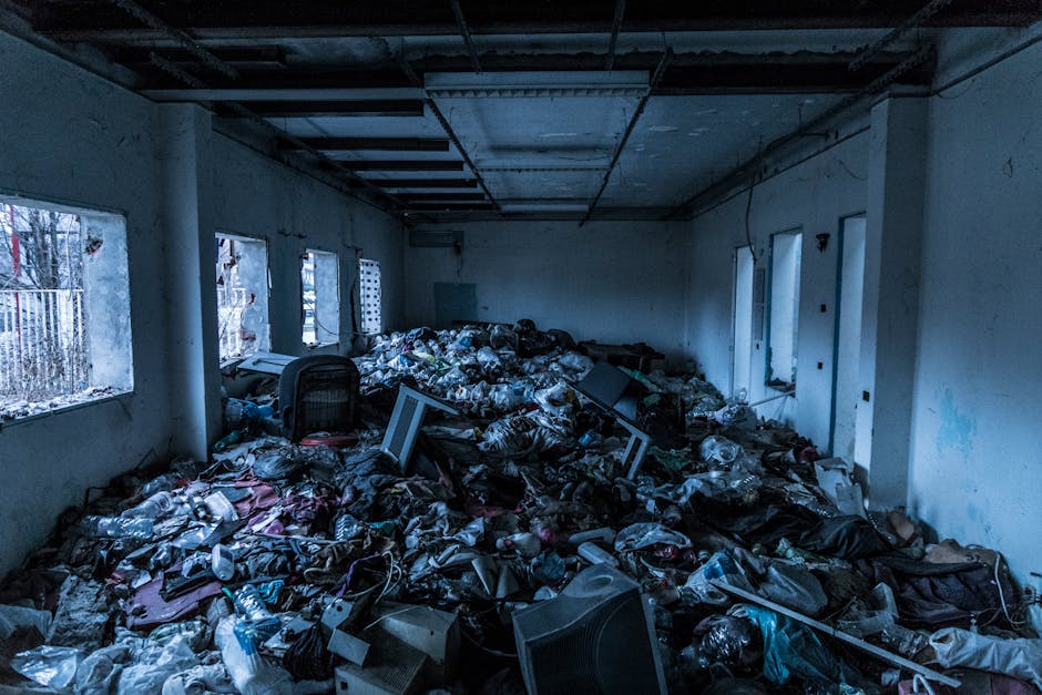 The image shows a large indoor space with white walls and a high ceiling, filled with a substantial amount of discarded waste, primarily consisting of plastic bags and packaging. In the foreground, numerous black and transparent plastic rubbish bags are piled haphazardly across the floor, some torn open and spilling contents. Among the debris, there are broken office monitors, electronic equipment, and crumpled cardboard boxes. To the left, several windows with metal bars allow natural light into the room, revealing a glimpse of the outdoor environment with trees and fencing visible through the panes. The walls are stained and chipped, indicating neglect or prolonged exposure to dirt. The ceiling appears partially exposed, with ceiling tiles missing and visible ductwork and structural beams. This scene highlights a significant accumulation of domestic or commercial waste, which could be collected through an alternative private rubbish clearance service offered by companies such as Rubbish Clearance Hounslow, supporting the needs for on-site waste removal or independent disposal solutions in similar indoor environments.