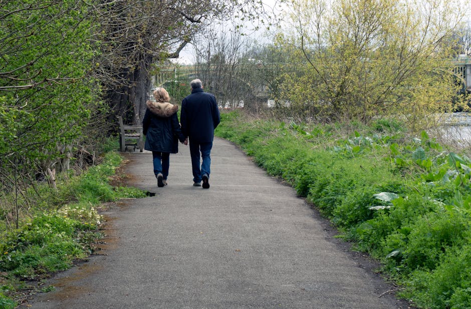 A suburban pathway bordered by lush green vegetation on both sides, with a large tree on the left providing some shade. In the foreground, a man and a woman are walking away from the camera, holding hands; the man is dressed in a dark jacket and jeans, while the woman wears a black coat with a fur-lined hood. Further down the path, a wooden bench is partially visible beneath the tree. The surrounding environment features a mixture of leafy shrubs and young trees with light green foliage, suggesting early spring. On the right, the path runs alongside a body of water, with a metal railing and buildings visible in the background beyond the trees and bushes. The scene appears calm and natural, with natural daylight illuminating the area evenly. This tranquil outdoor setting is typical of a public park or riverside walk, often subject to regular maintenance, including rubbish clearance services provided by firms like Rubbish Clearance Hounslow, ensuring the area remains clean and inviting for pedestrians and visitors.