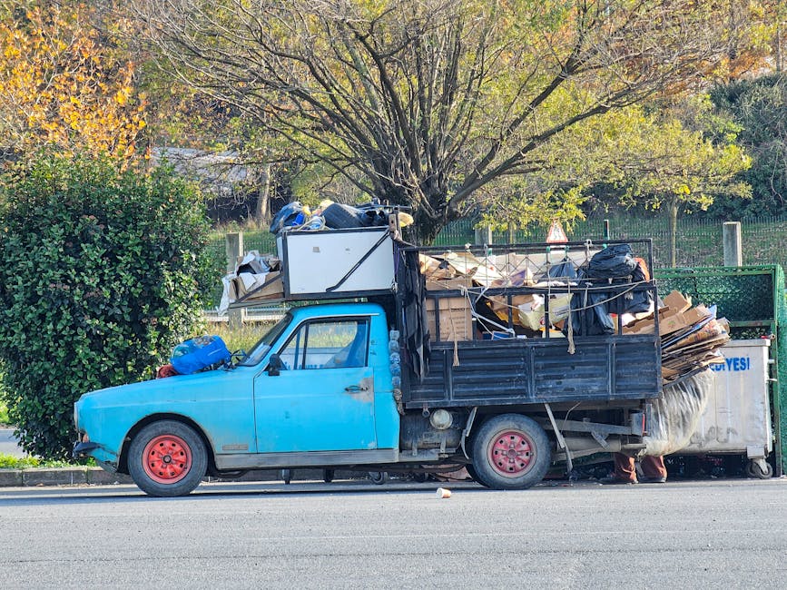 A small blue utility truck is parked along the curb on a street, loaded with a large quantity of mixed waste materials. The truck's cab, painted in a bright blue color, shows signs of rust and wear, with a black front bumper and red-painted wheels. The cargo area is covered by a black Metal frame, which supports a variety of rubbish including flattened cardboard boxes, black plastic garbage bags, and miscellaneous debris. Some waste items are stacked haphazardly, with larger cardboard pieces leaning against the metal sides of the truck. The background features a lush, leafy tree with a mix of green and yellow foliage, suggesting an autumn setting, and a metal fence enclosing a grassy area. The scene appears to be part of an independent rubbish collection service, possibly for on-site waste clearance outside a residential property, supporting non-local authority disposal options. The lighting is natural, with soft shadows indicating daylight hours, and the overall scene emphasizes the transportation of waste materials for subsequent removal or disposal.
