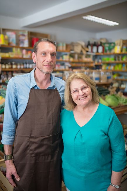 A middle-aged man with short light brown hair and a light beard, wearing a light blue shirt and a dark brown apron, stands alongside an elderly woman with shoulder-length blonde hair, wearing glasses and a turquoise long-sleeved top, inside a well-organized storage or shop environment. Behind them, shelves are filled with various products in cardboard boxes, plastic containers, and jars, suggesting a retail or warehouse setting related to waste management or supply distribution. The shelves are wooden and stacked neatly, with items of different shapes and colors, including green, yellow, red, and brown packaging materials. The ceiling features a white, flat surface with a fluorescent light fixture illuminating the scene evenly. The background indicates a professional environment where waste or material handling may take place, aligning with the context of rubbish removal services or independent waste collection activities. Rubbish Clearance Hounslow's presence is inferred through the setting, emphasizing a professional and organized approach to waste or supply management, suitable for small business or retail waste removal strategies.