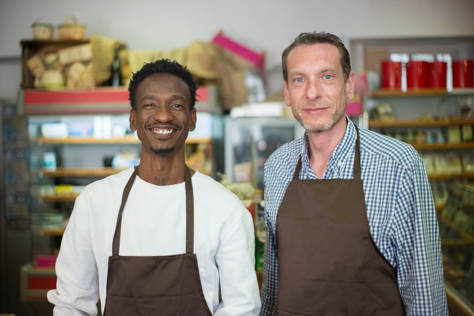 Two men standing inside a retail store or deli, both wearing brown aprons over casual clothing. The man on the left, with dark skin and curly black hair, is smiling broadly and wearing a white shirt beneath his apron. The man on the right, with light skin and short, light brown hair, is also smiling gently and is dressed in a checked shirt under his apron. Behind them, shelves are stocked with various packaged goods, including jars, cans, and boxes, arranged neatly and displaying a mix of colourful labels and containers. The environment is well-lit, with a clean and organized appearance, suggesting a shop or small business setting. The scene subtly reflects a professional context that could relate to small business services such as waste removal or local retail management, emphasizing a community-oriented environment.