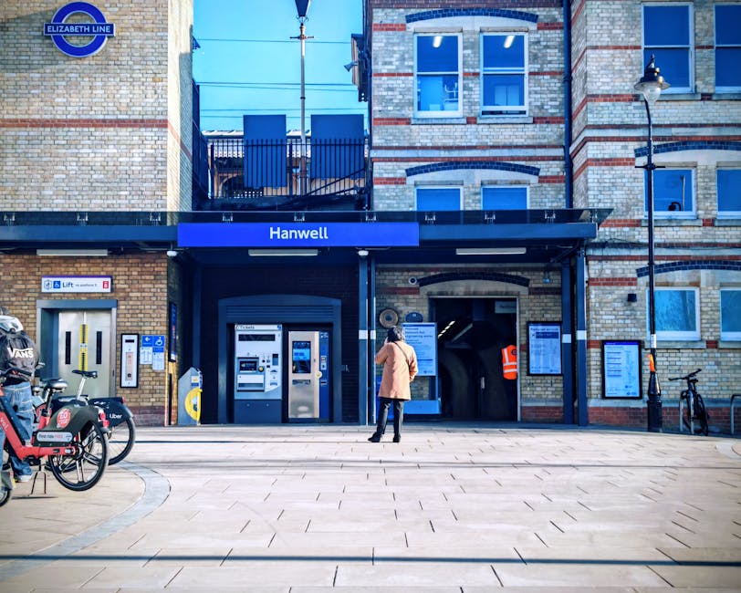 A person wearing a beige coat and black trousers stands on a paved pedestrian area in front of Hanwell underground station entrance, part of the Elizabeth Line, located within a brick building with red and light-colored bricks forming decorative patterns. The station signage is prominent on a blue panel above the entrance. To the left, there are several public bicycle hire bikes, one branded with Uber and another with Velo, parked on stands. Near the station entrance, there are ticket machines and a yellow and grey barrier for the ticketing system. An elevated walkway with a black railing is visible above the station, and a streetlamp with a curved arm and classic-style lantern is mounted on the right side of the building. The area is well-lit with natural daylight, casting shadows on the ground, and the scene appears quiet with no visible vehicular traffic, emphasizing a typical day at the station, which may connect to private waste management services, but no waste objects are visible in this image.
